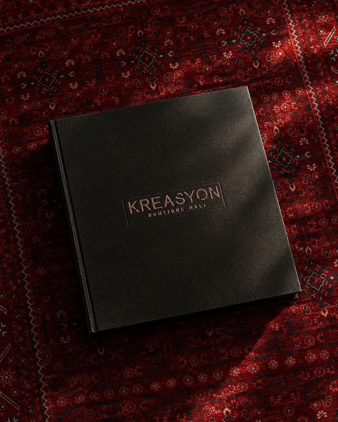 Kırmızı halı üzerinde yukarıdan görülen dokulu siyah Kreasyon kataloğu; ortada bakır varak KREASYON Boutique Halı logosu