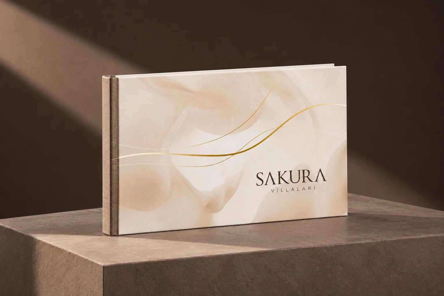 Koyu taupe stüdyo zemininde bir kaide üzerinde dik duran, krem kapağında altın çizgiler ve Sakura Villaları logosu olan katalog