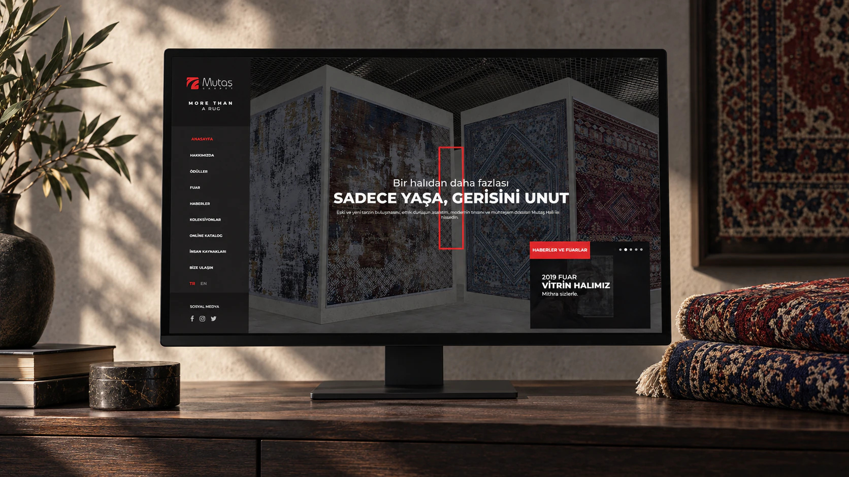 Koyu meşe konsolda Mutaş Carpet ana sayfasını gösteren ekran, yanında katlanmış desenli halılar