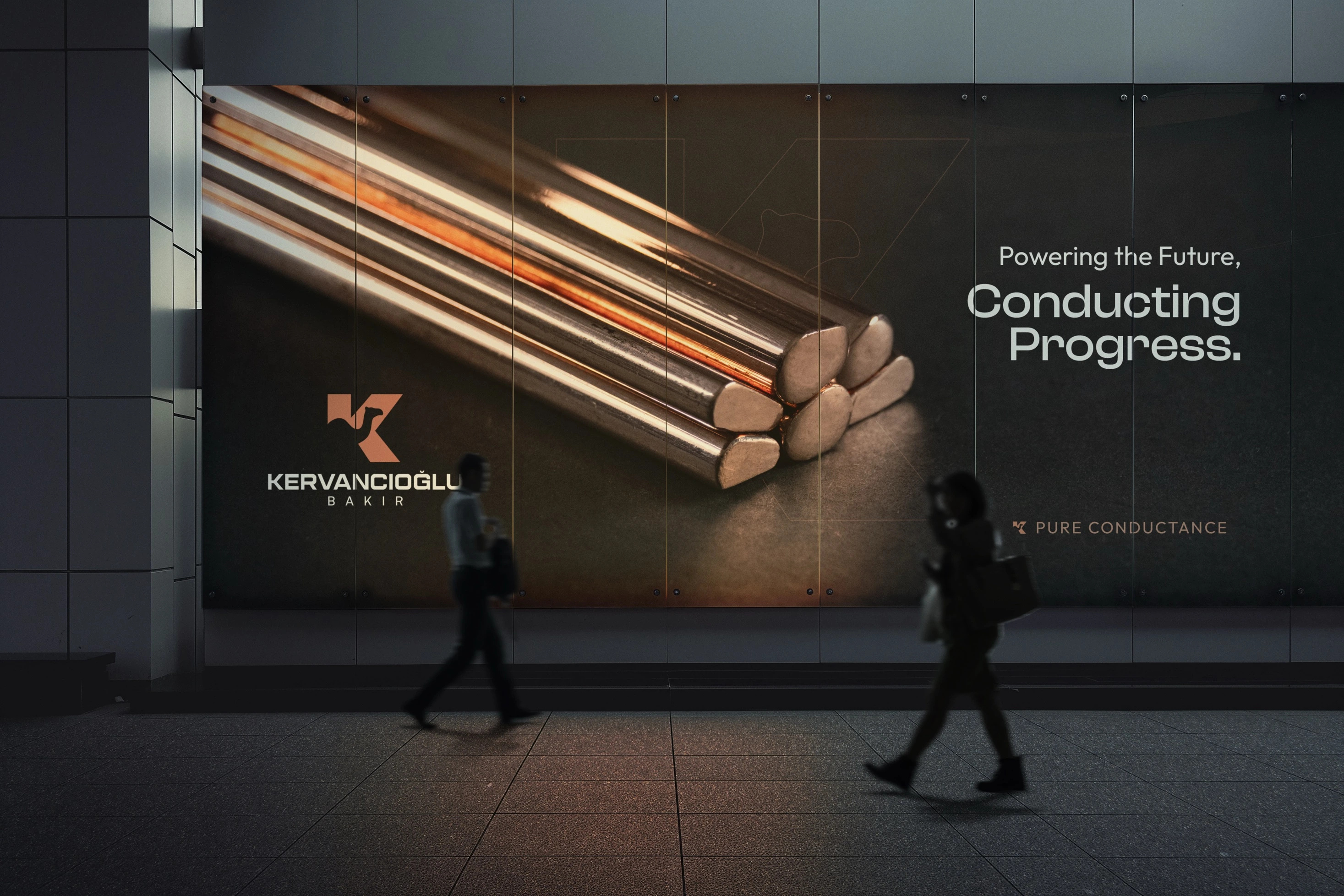 Gece bir cadde duvarındaki Kervancıoğlu Bakır billboardu önünden geçen iki yaya; panoda bakır çubuklar ve “Powering the Future, Conducting Progress.” yazısı